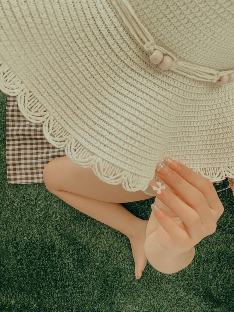 Na obrazku widać osobę siedzącą na trawie w słomkowym kapeluszu z szerokim rondem i frędzlami. Kapelusz ma naturalny, kremowy kolor i ozdobne sznurki z kulkami. Osoba ma na sobie wzorzystą bluzkę w kratę w kolorach brązowym i białym. Na dłoniach widać delikatne, białe naklejki lub tatuaże w kształcie kwiatków. Całość ma ciepłą, letnią atmosferę z miękkim, naturalnym oświetleniem.